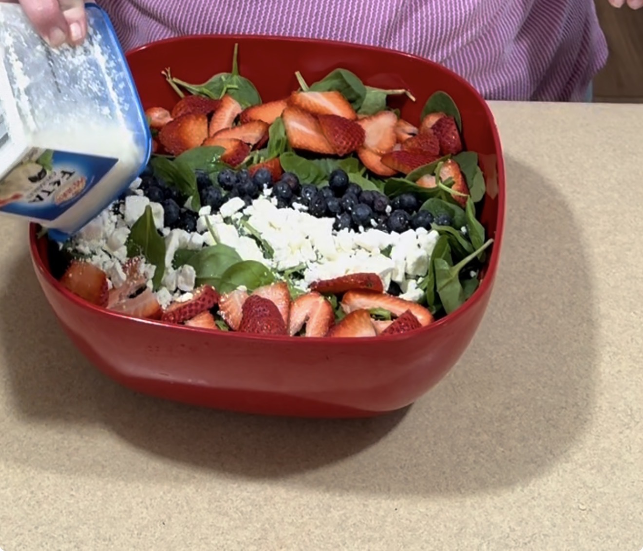 A person pours salad dressing from a bottle onto a spinach salad topped with sliced strawberries, blueberries, and crumbled cheese in a red bowl on a countertop.