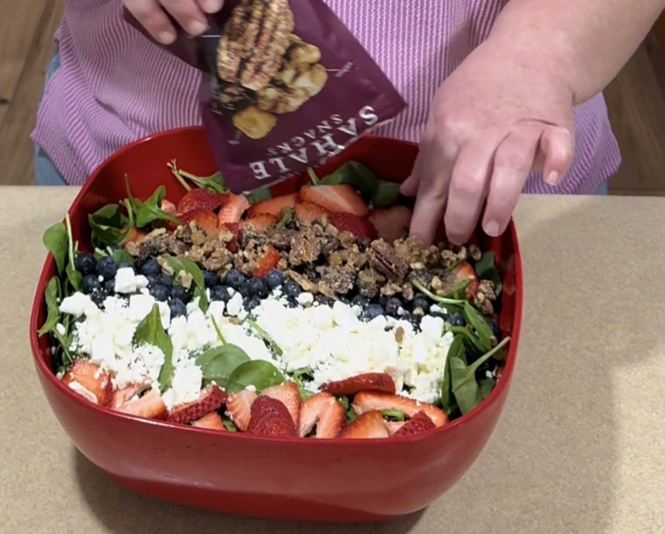 A person sprinkles packaged granola onto a fresh salad in a red container. The salad contains spinach, blueberries, sliced strawberries, and crumbled feta cheese.