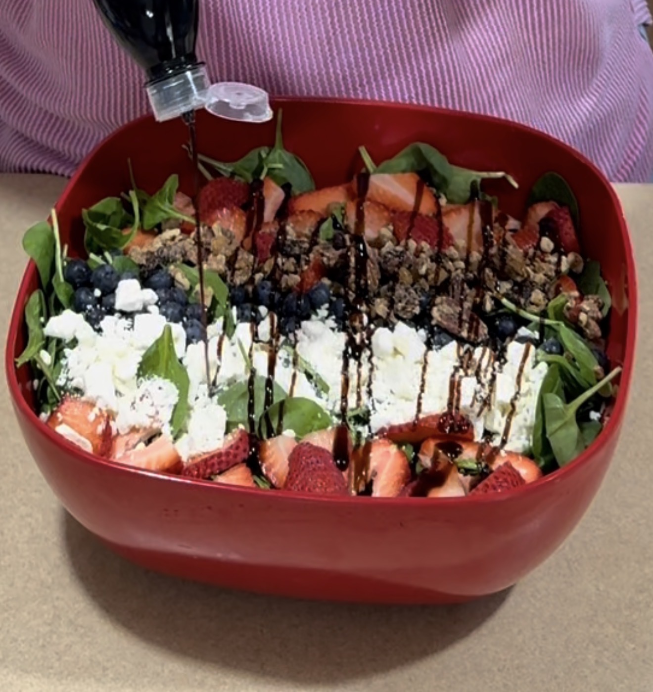 A person pours balsamic glaze onto a salad in a red bowl, topped with spinach, sliced strawberries, blueberries, feta cheese, nuts, and granola.