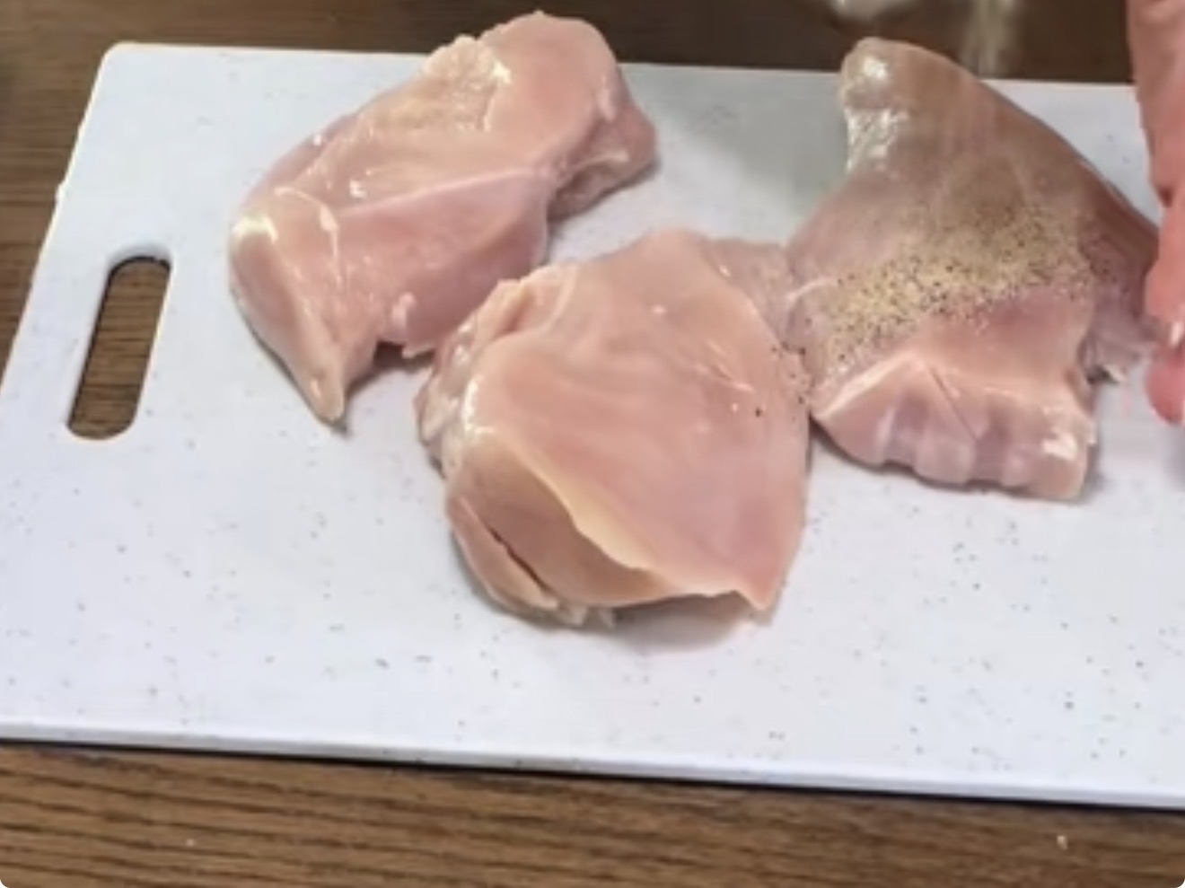 Three raw chicken breasts on a white cutting board; a hand is sprinkling seasoning, likely salt and pepper, onto the chicken.