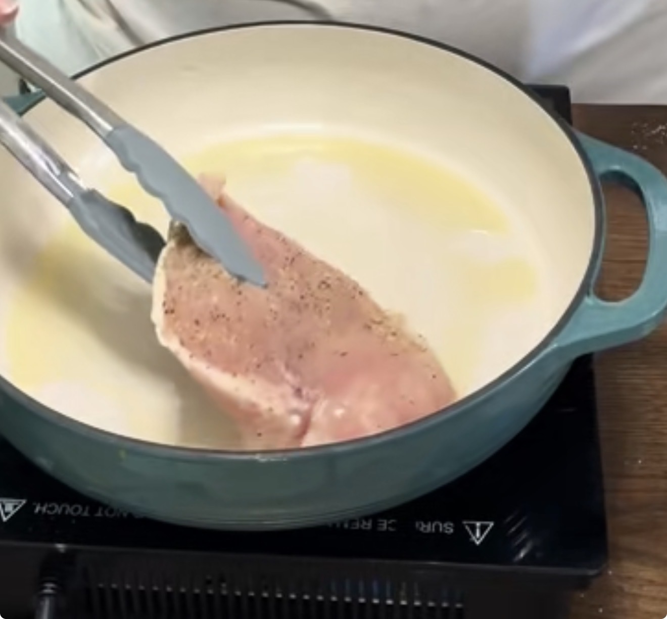 A person holds raw, seasoned chicken breast with metal tongs over a turquoise pot on an electric stovetop, preparing to cook it in melted butter.