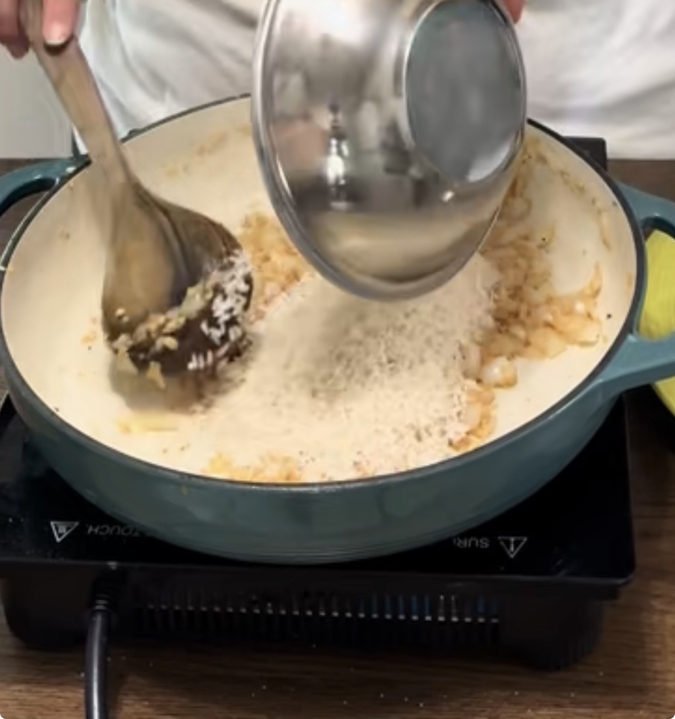A person pours uncooked rice from a metal bowl into a pot with sautéed onions, stirring with a wooden spoon on an electric stovetop.