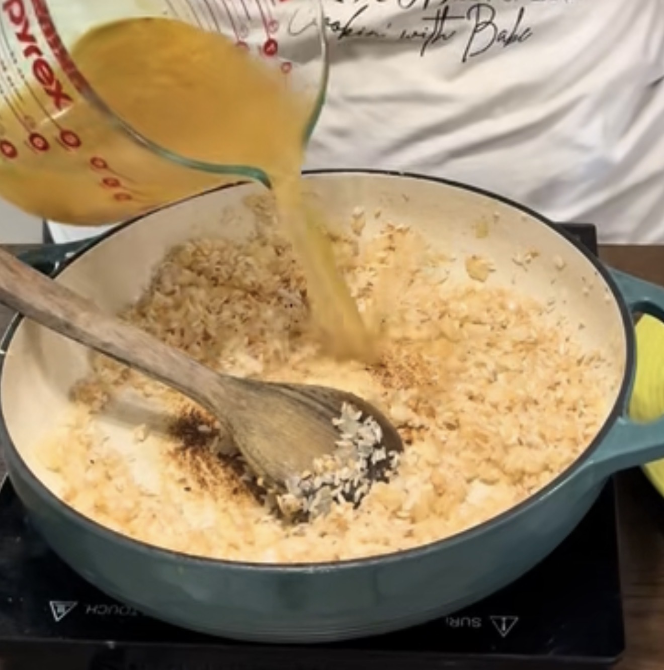 A person pours broth from a glass measuring cup into a pot of sautéed onions and rice, stirring with a wooden spoon on a stovetop.
