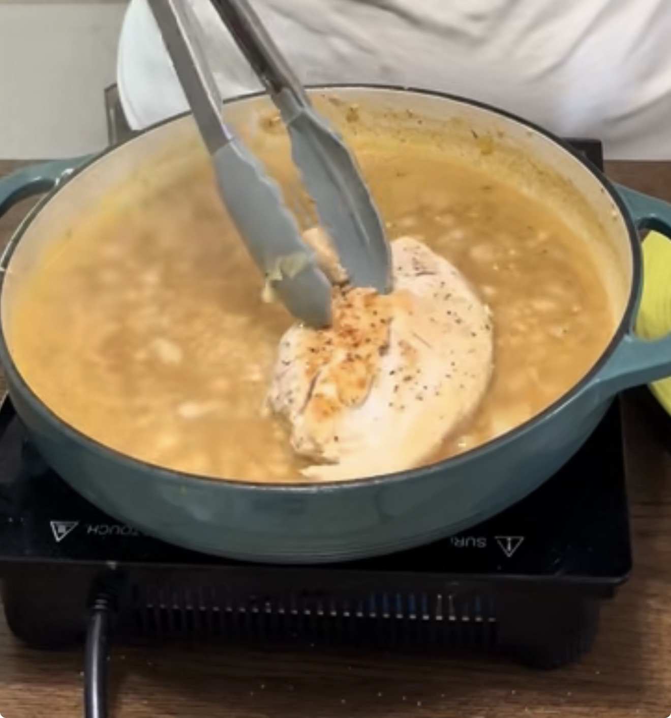 A person uses metal tongs to hold a seasoned chicken breast cooking in a round green pot filled with simmering broth on an electric stovetop.