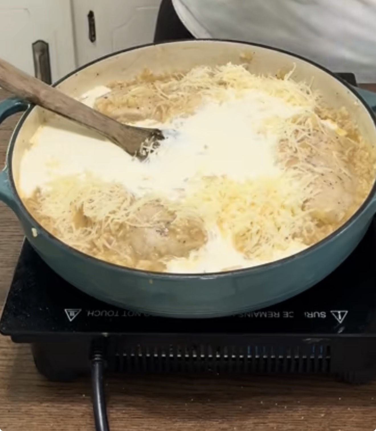 A round green pot on a hot plate contains chicken in a creamy sauce topped with shredded cheese, being stirred with a wooden spoon. The scene is set on a wooden countertop.