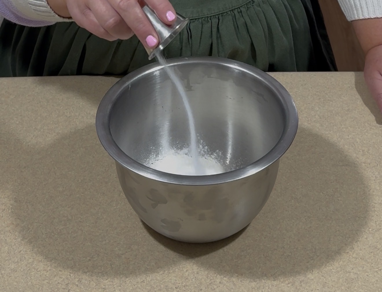 A person pours sugar from a small metal cup into a stainless steel mixing bowl containing white powder, likely flour, on a kitchen counter.