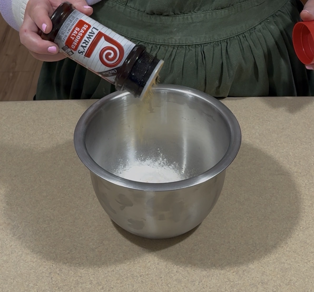 A person pours Lawry’s seasoned salt from a bottle into a metal mixing bowl containing a white powder, on a kitchen counter.