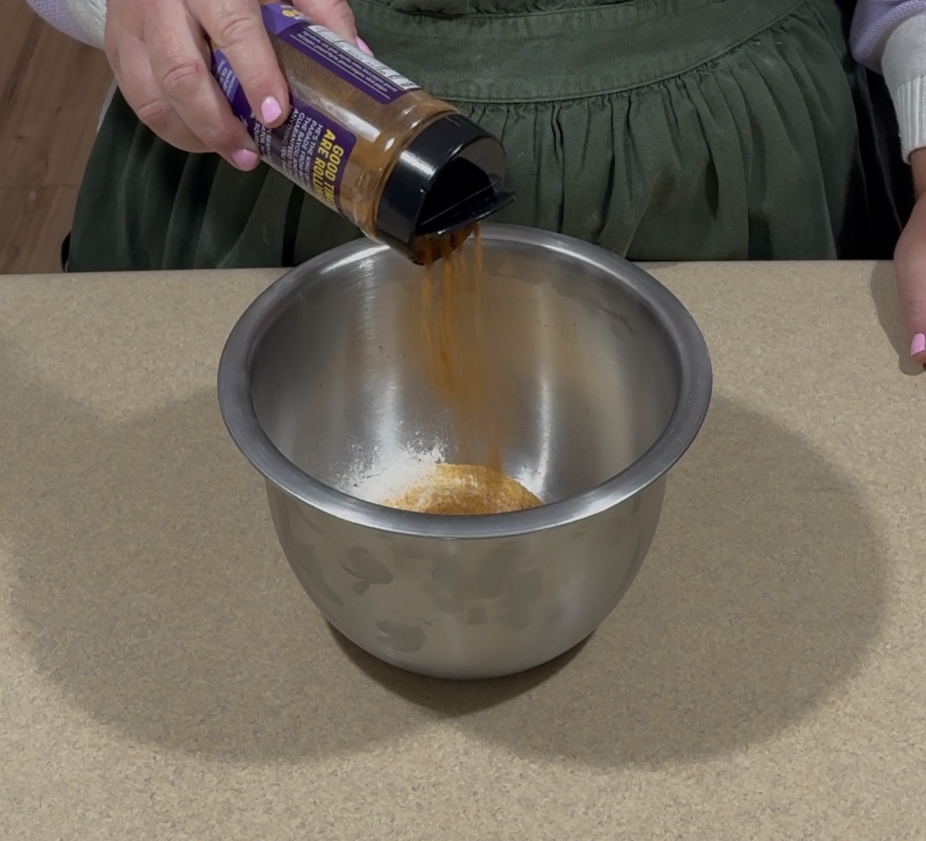 A person pours seasoning from a spice container into a metal mixing bowl on a countertop.