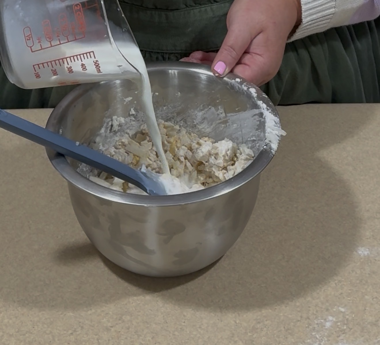 A person pours milk from a measuring cup into a mixing bowl filled with flour and chopped ingredients, using a blue spatula to mix, on a kitchen countertop.