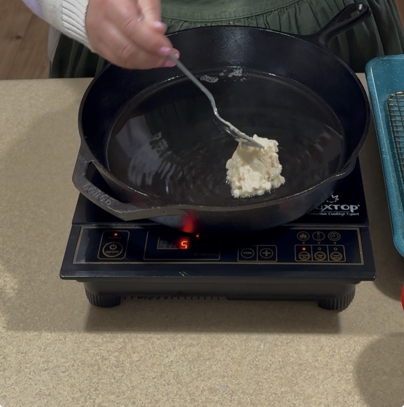 A person uses a spoon to drop batter into a black cast iron skillet with oil, which is heating on an electric stovetop set to level 5. A blue cooling rack sits on the counter nearby.