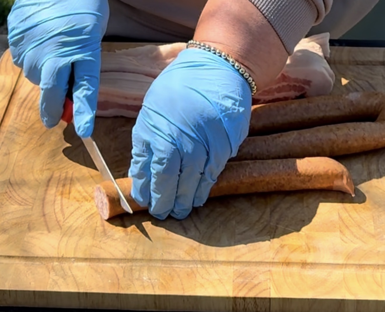A person wearing blue gloves slices sausage on a wooden cutting board with a knife. Other sausages and a piece of raw meat are also on the board.