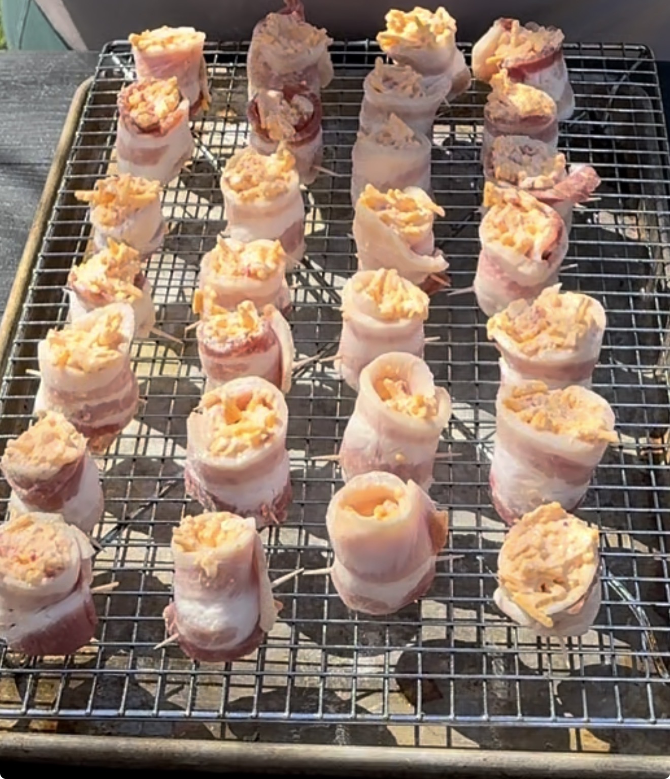 Rows of bacon-wrapped, cheese-stuffed rolls are arranged on a wire rack placed over a baking sheet, ready to be cooked in the sunlight.