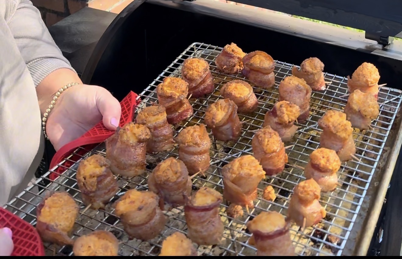 A person uses red silicone grips to hold a metal rack filled with rows of bacon-wrapped appetizers, fresh from a grill or smoker. The bite-sized snacks appear golden and crispy.