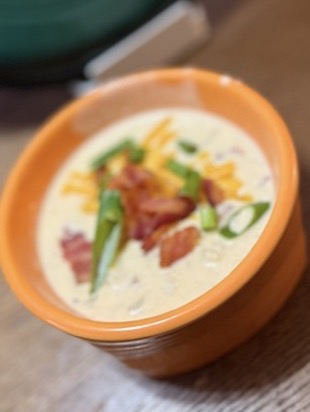 A bowl of creamy soup topped with shredded cheese, bacon pieces, and chopped green onions, served in an orange bowl on a wooden table.
