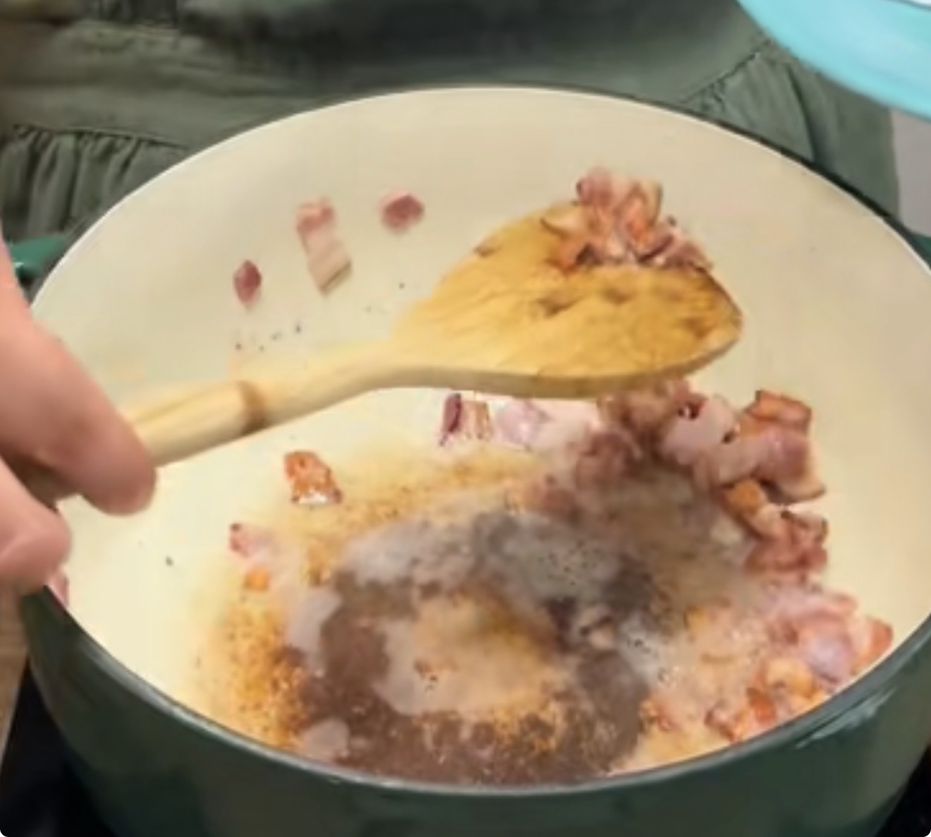 A person stirring chopped bacon pieces in a large white pot with a wooden spoon, as the bacon cooks and browns on the bottom of the pot.