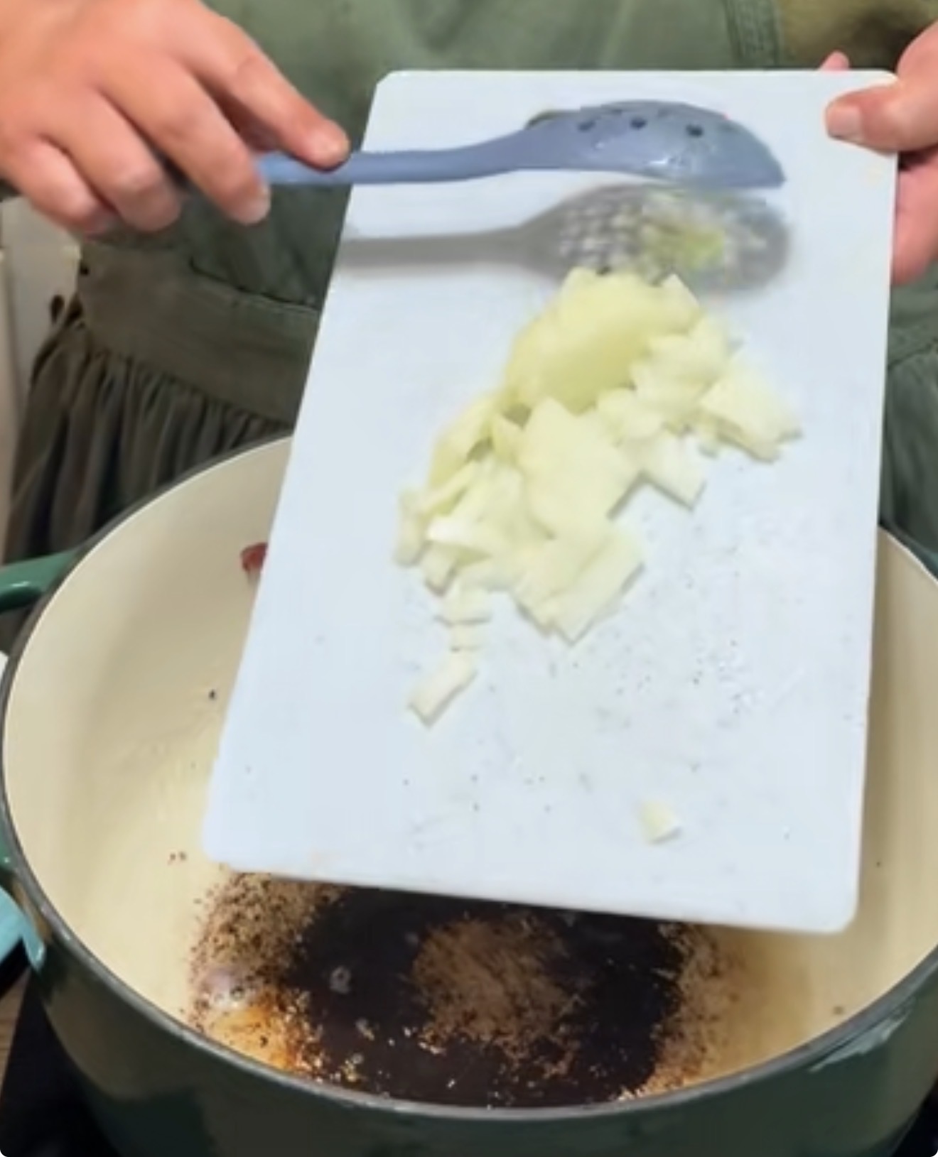 A person pours chopped onions from a white cutting board into a pot on a stove, using a blue slotted spoon to help guide the onions.