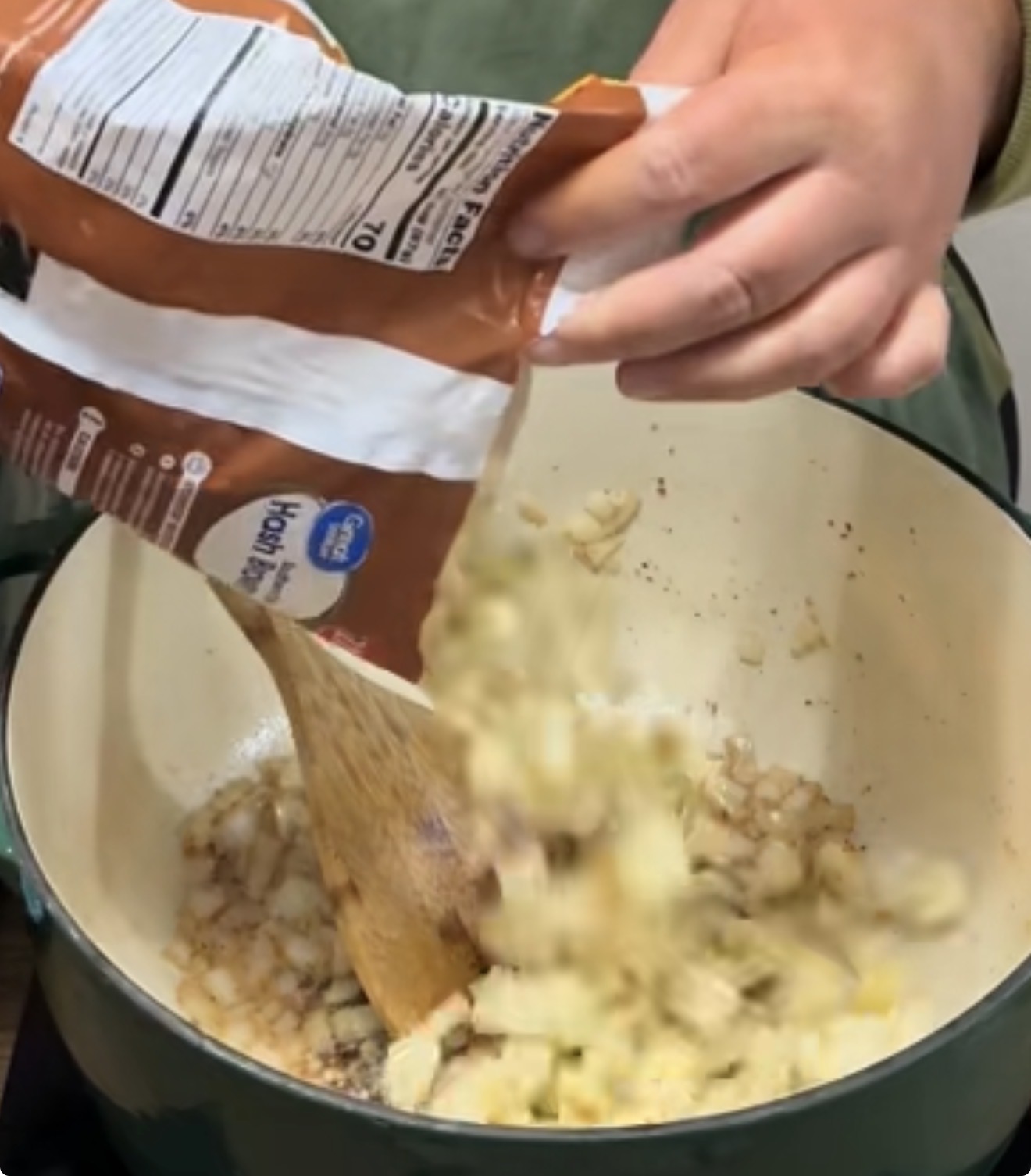 A person pours salt from a brown bag into a pot of chopped onions, stirring with a wooden spoon.