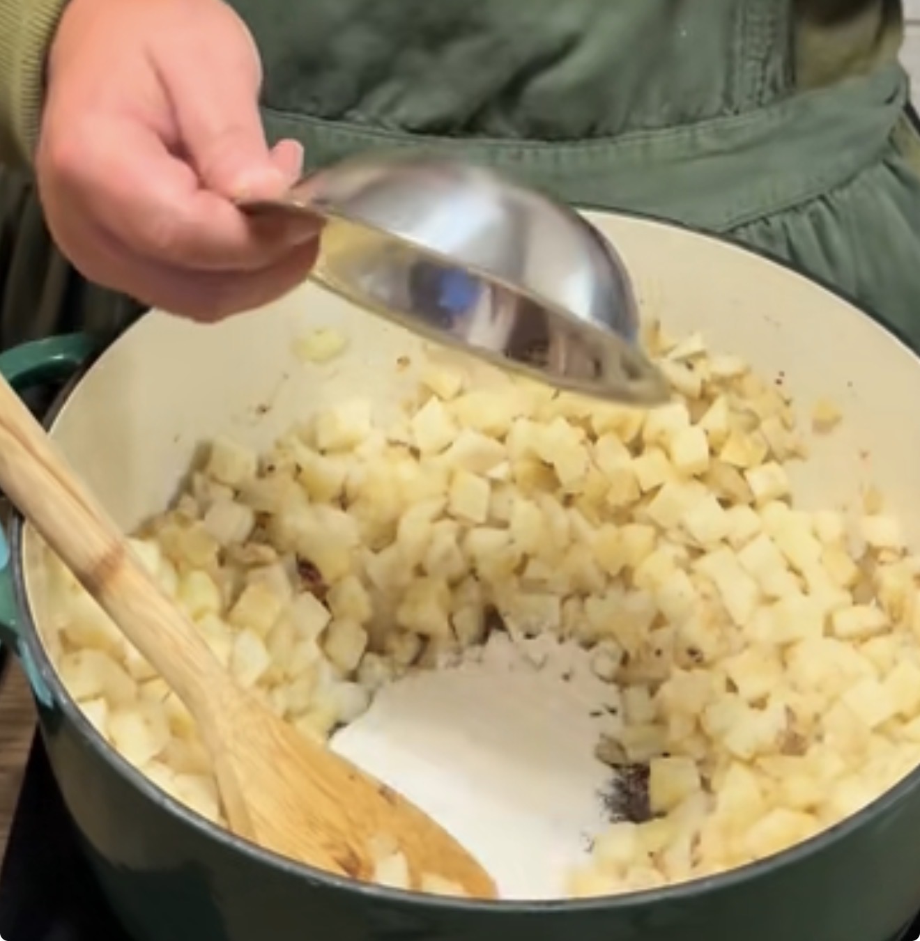 A person in a green apron pours sugar from a metal bowl into a pot of diced apples, stirring with a wooden spoon.