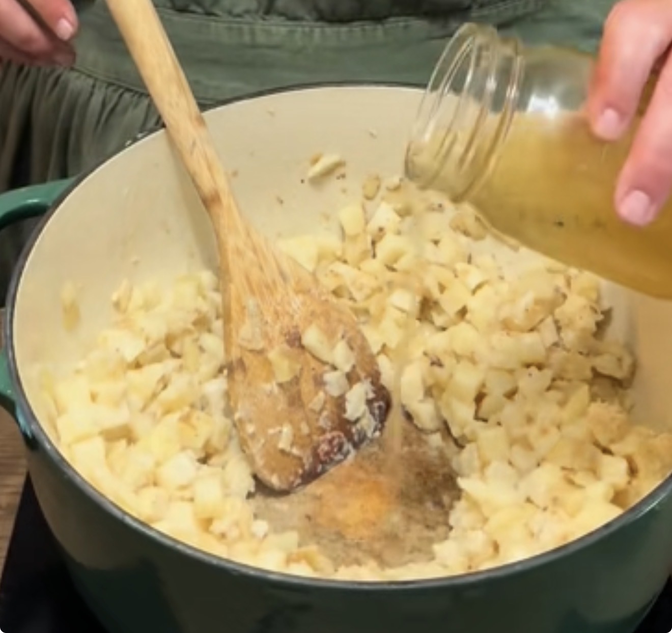 A hand pours broth from a jar into a pot filled with diced potatoes, while stirring with a wooden spoon. The scene suggests the beginning steps of making soup or stew.