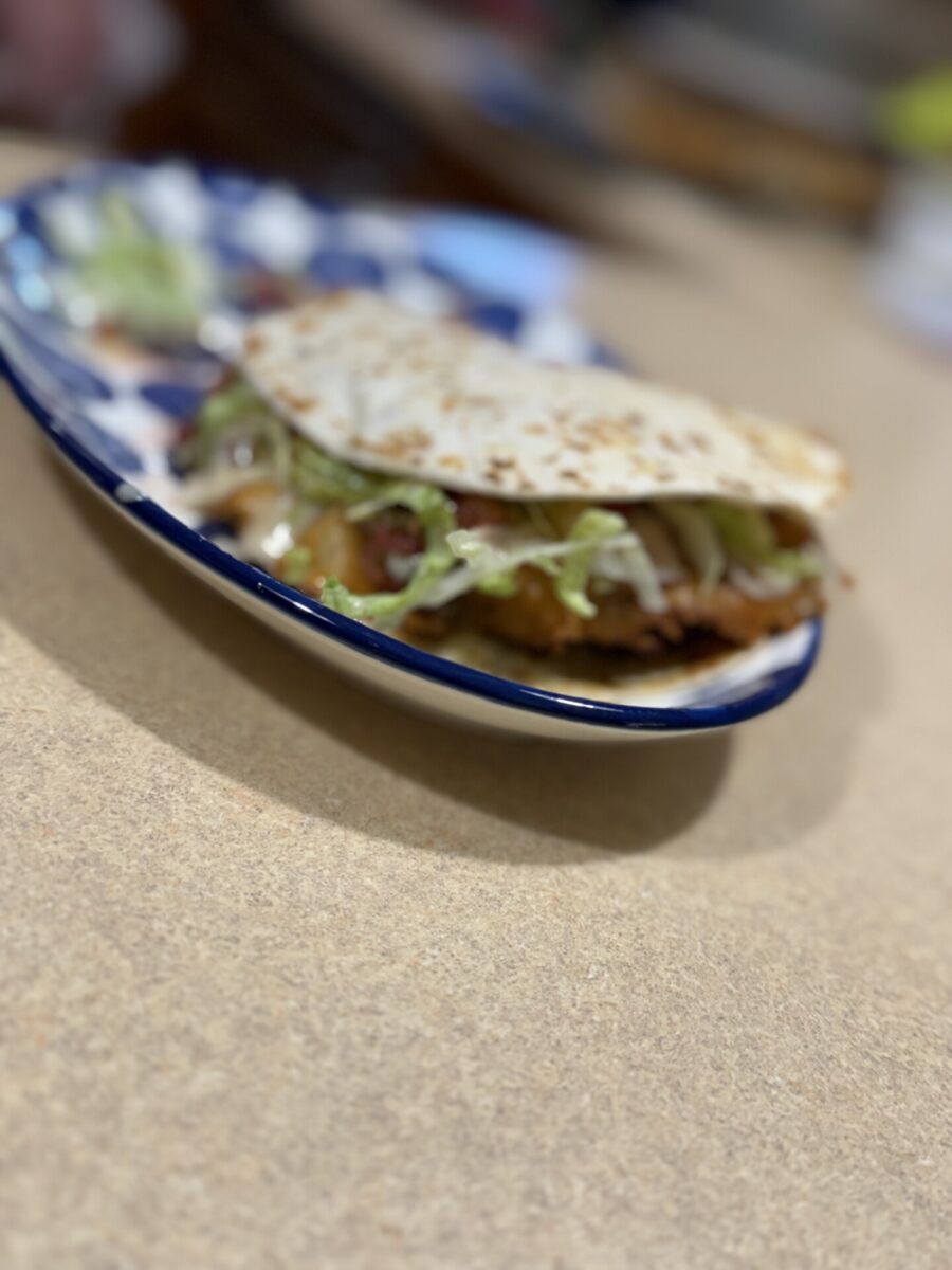 A close-up of a taco with lettuce, cheese, and other fillings on a blue and white plate, placed on a light brown countertop. The background is blurred.