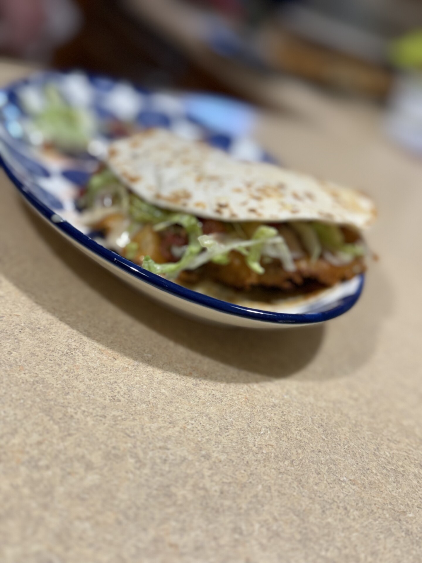 A close-up of a taco with lettuce, cheese, and other fillings on a blue and white plate, placed on a light brown countertop. The background is blurred.