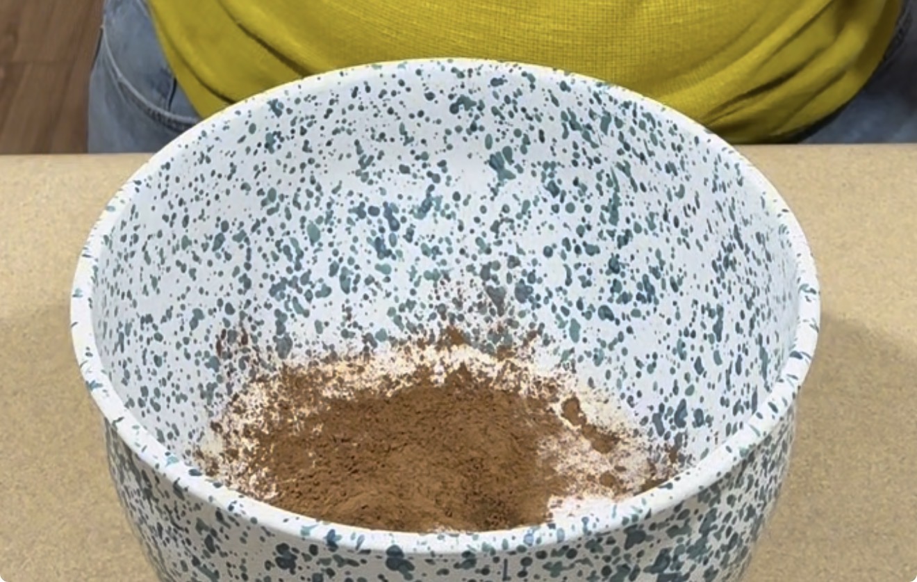 A speckled white and blue mixing bowl containing a pile of cocoa powder and flour sits on a countertop, with a person in a yellow shirt visible behind it.