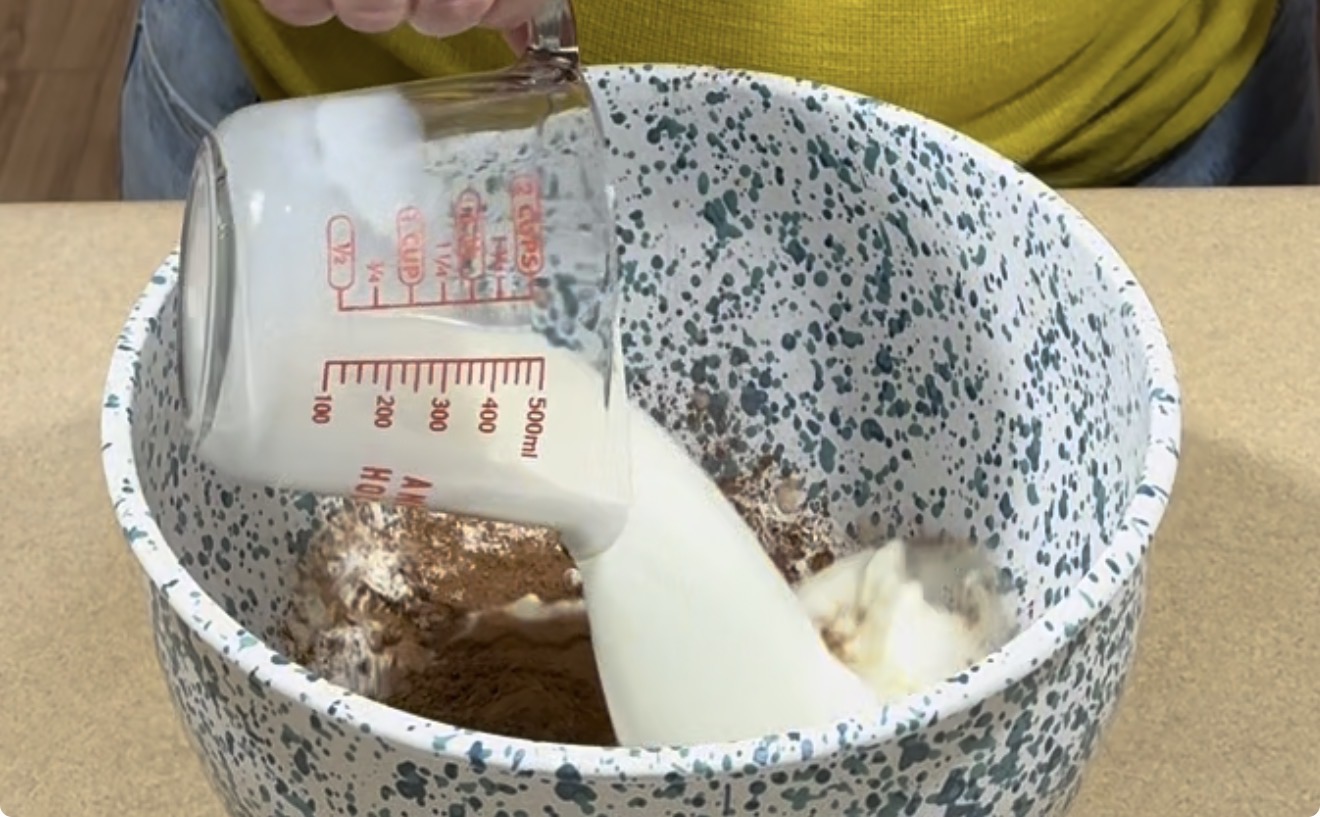 A person pours milk from a measuring cup into a speckled mixing bowl containing dry ingredients, likely for baking. The person is wearing a yellow shirt.