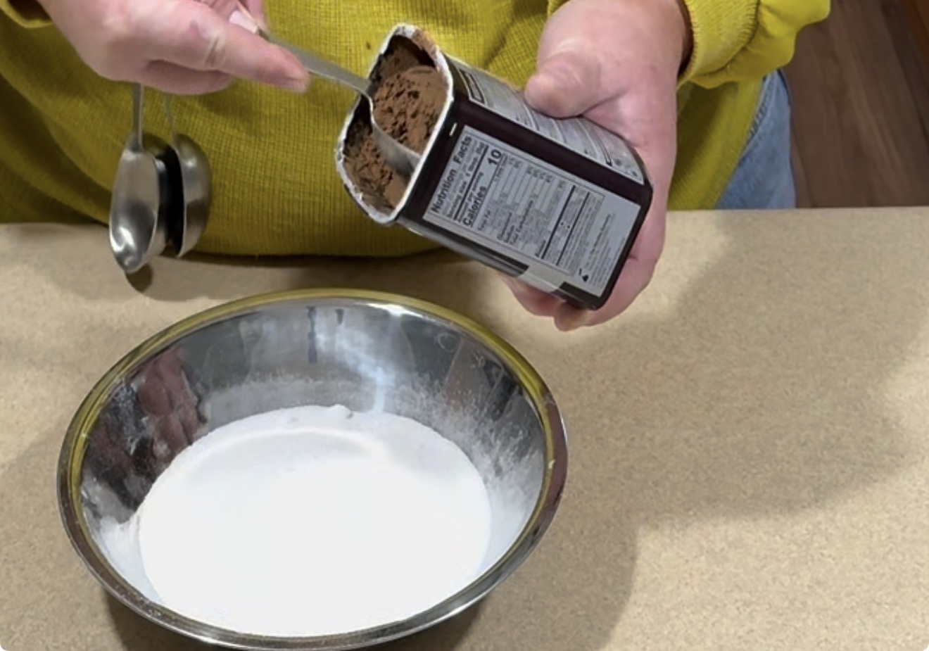 A person in a yellow sweater scoops cocoa powder from a container over a metal bowl filled with a white powder, possibly flour or sugar, on a beige countertop.