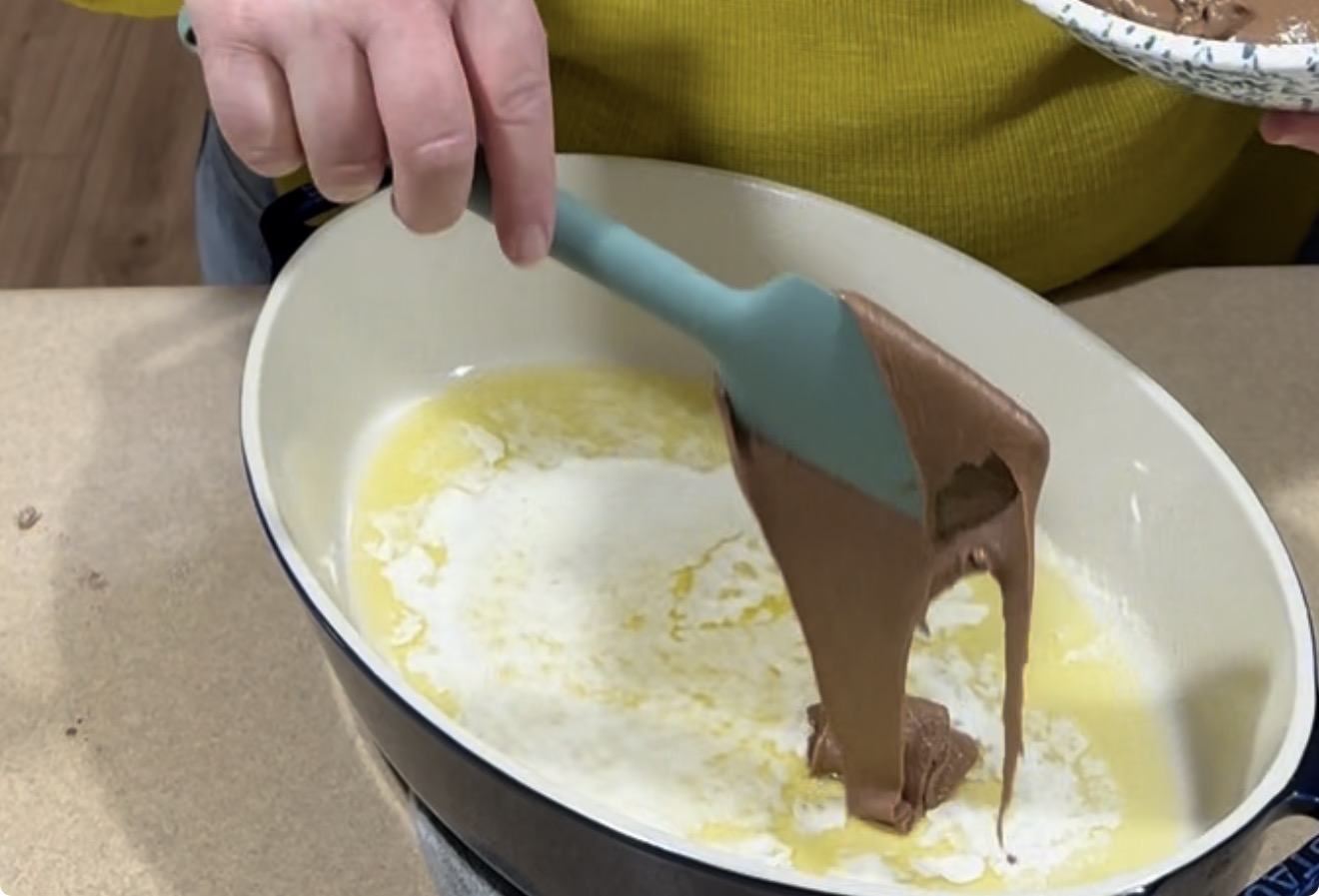 A person in a yellow shirt uses a teal spatula to spread chocolate batter over melted butter in a white casserole dish on a countertop.