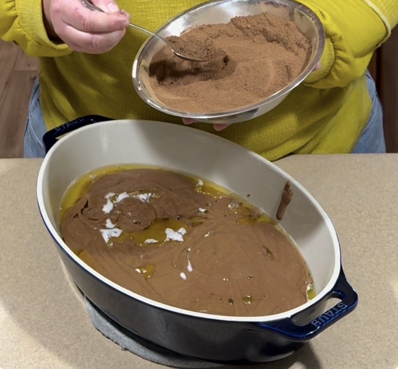 A person in a yellow shirt holds a bowl of cocoa powder over a casserole dish filled with a chocolate baking mixture, sprinkling the powder on top with a spoon.