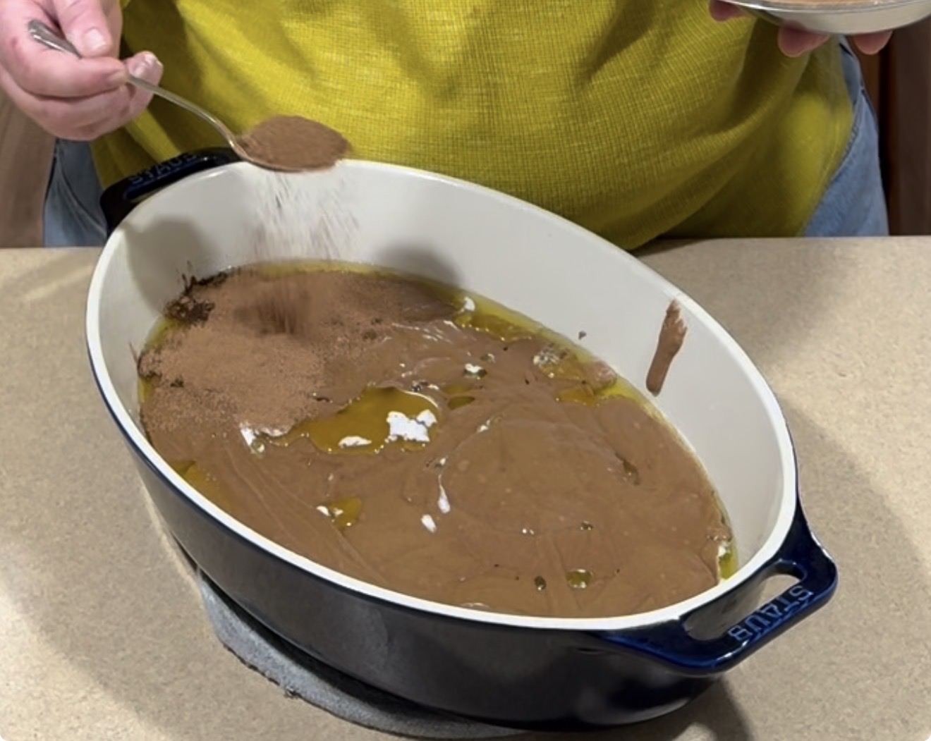 A person in a yellow shirt sprinkles cocoa powder from a spoon over a mixture of oil and batter in a large oval baking dish on a countertop.