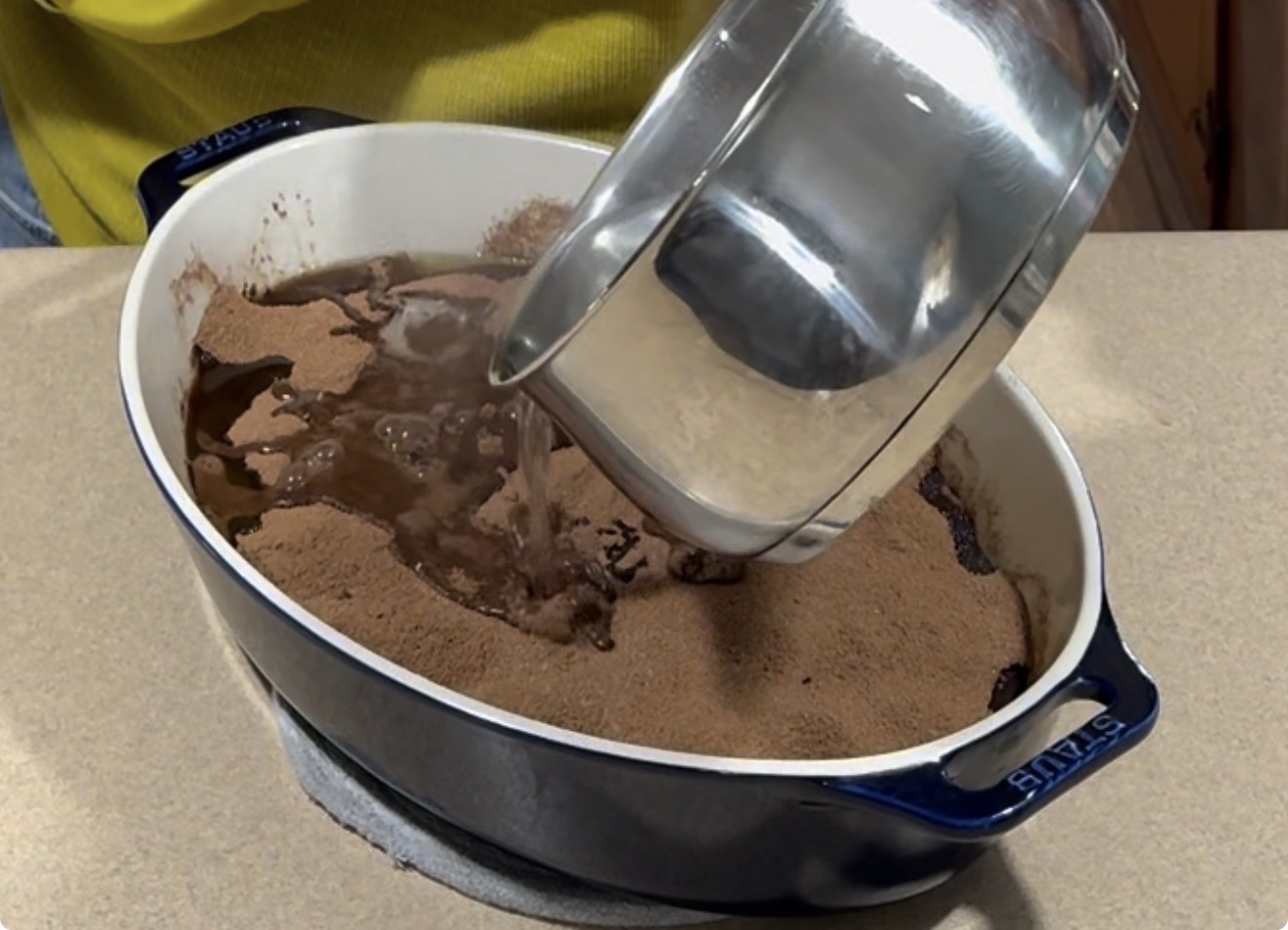 A person pours hot water from a metal pot onto a layer of cocoa powder and dry cake mix in a blue baking dish on a kitchen counter.