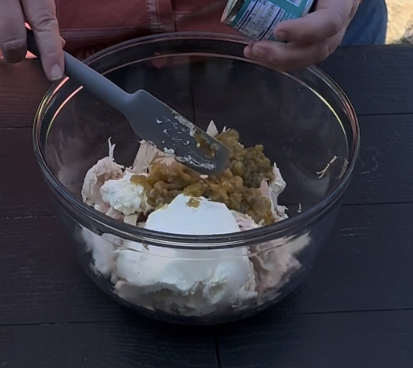 A person uses a spatula to combine canned green chiles, cream cheese, and shredded chicken in a clear glass mixing bowl on a dark table.