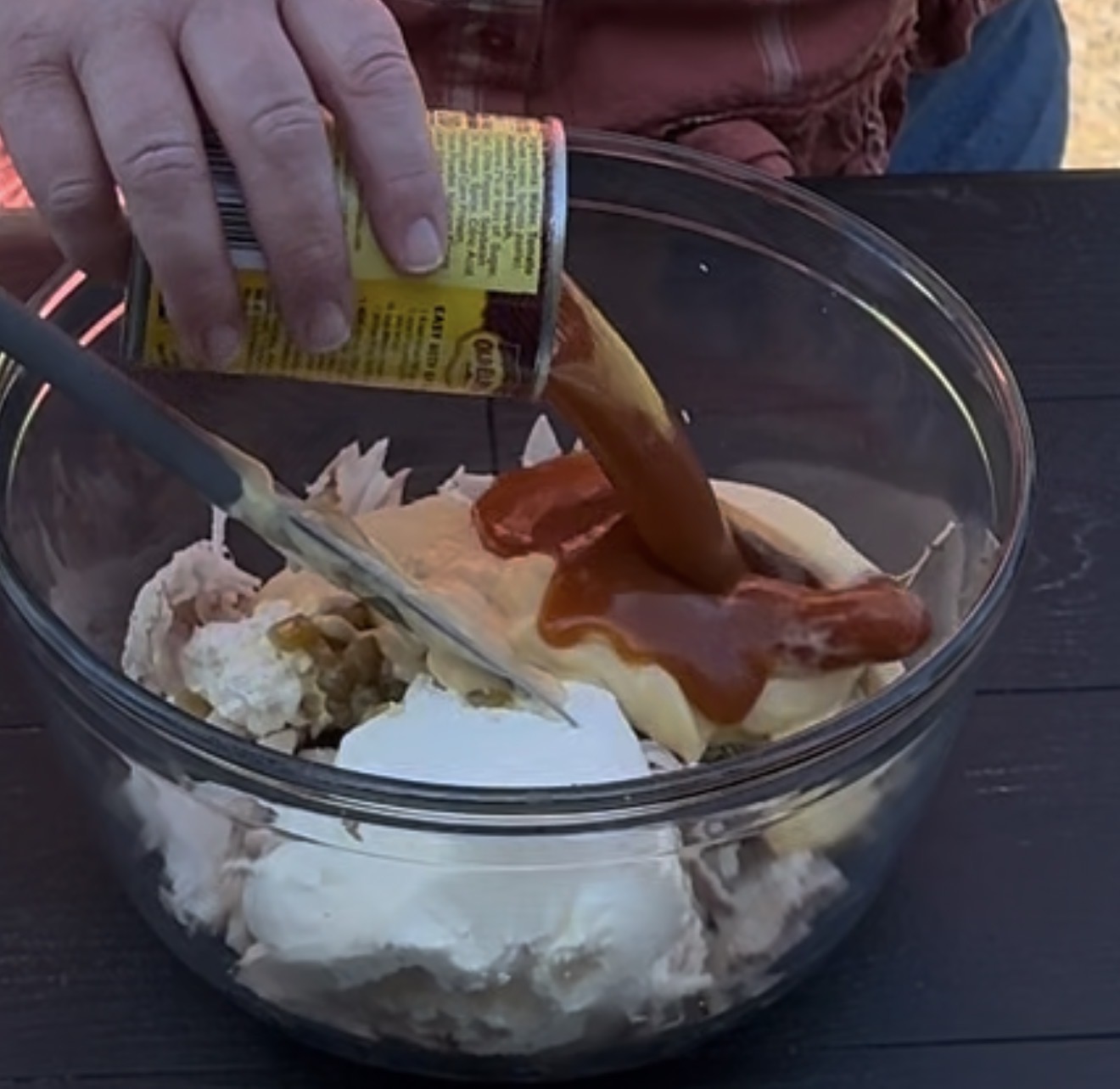 A person pours canned enchilada sauce into a glass bowl containing sour cream, cream cheese, shredded chicken, and other ingredients, while mixing them with a spatula.