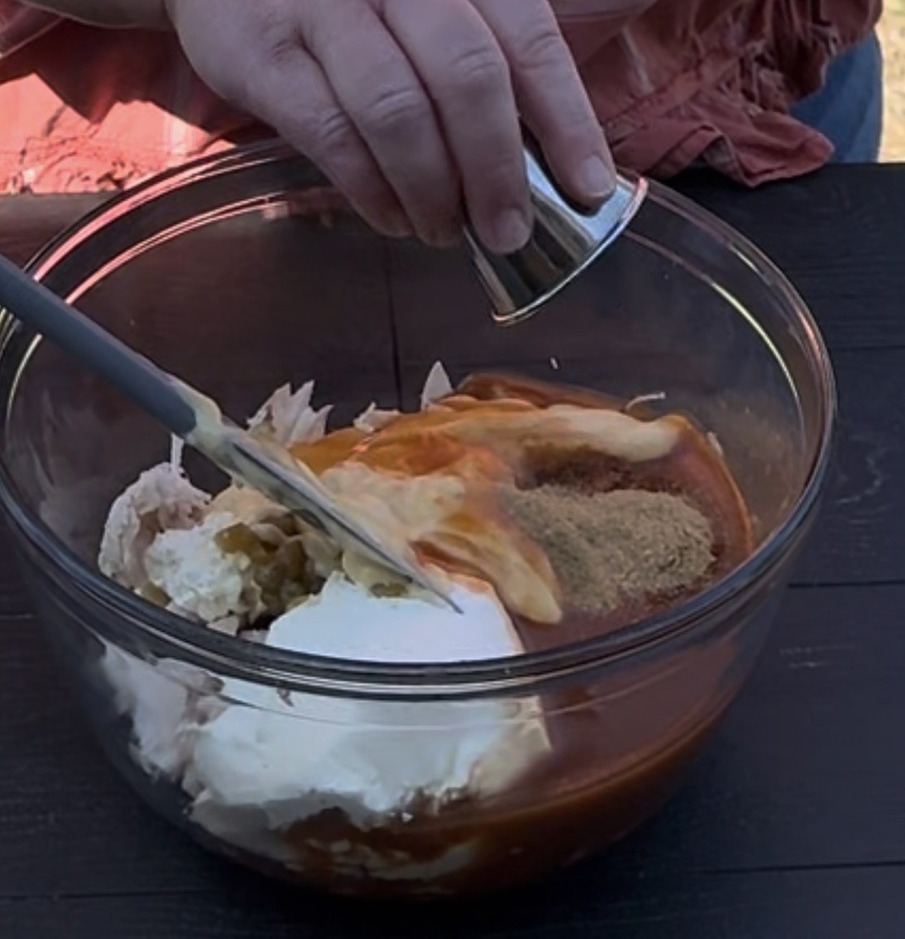 A person pours liquid from a small metal cup into a glass bowl filled with shredded chicken, sour cream, spices, and sauce, while holding a spatula.
