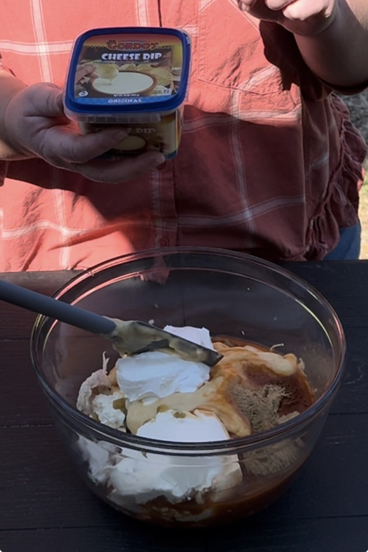 A person holding a container of Kraft Cheesy Dip stands next to a bowl filled with cream cheese, sour cream, and brown liquid ingredients being mixed with a spatula.