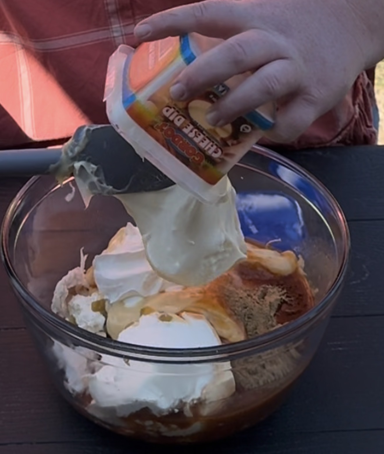 A person in a red shirt is adding creamy cheese dip from a container into a glass bowl filled with other ingredients for mixing.