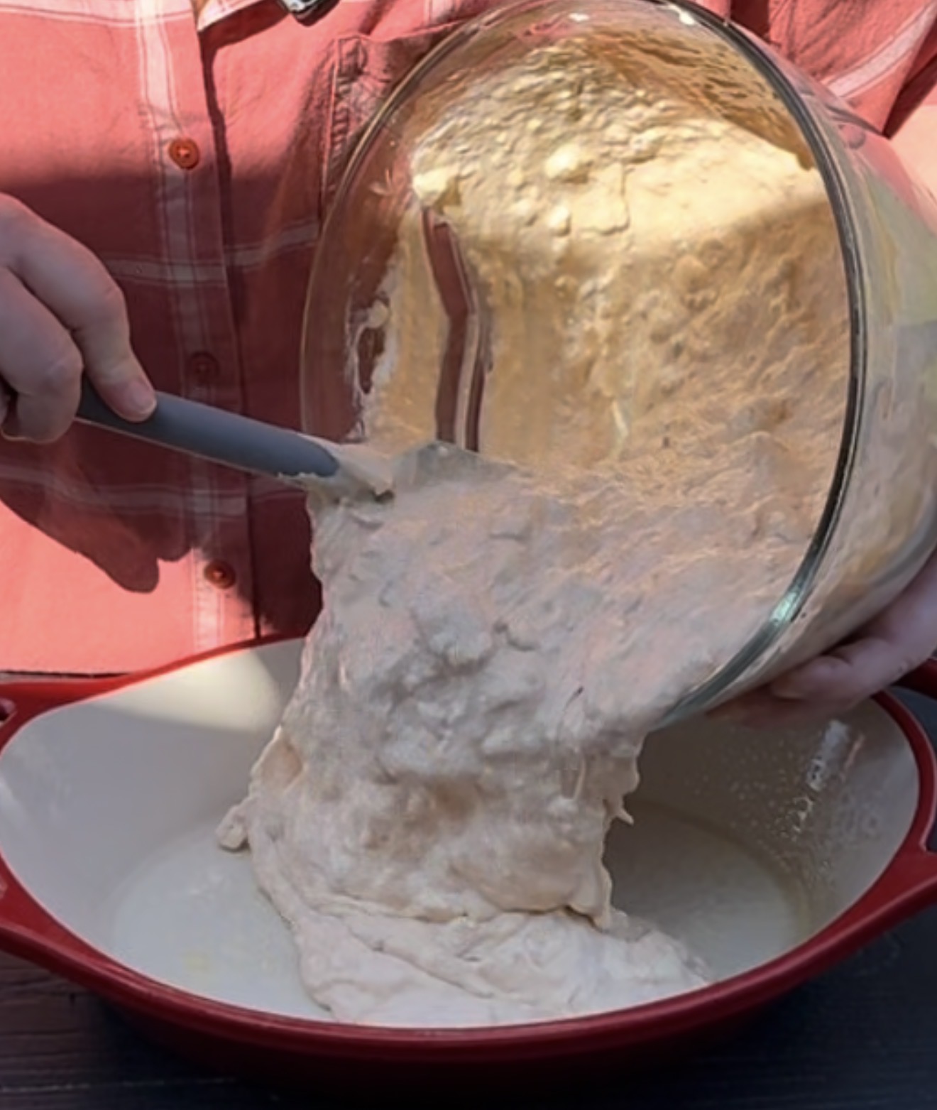 A person pours sticky bread dough from a large glass bowl into a red and white baking dish, using a spatula to help guide the dough.
