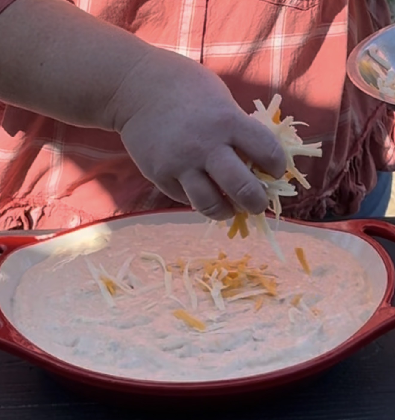 A person in a red plaid shirt sprinkles shredded cheese over an uncooked pizza crust in a red baking dish.