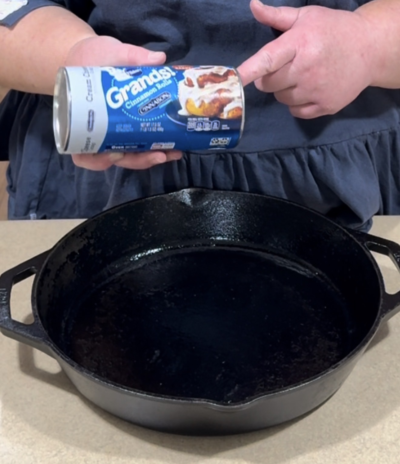 A person holds a can of Grands! cinnamon rolls above a black cast iron skillet on a kitchen counter.