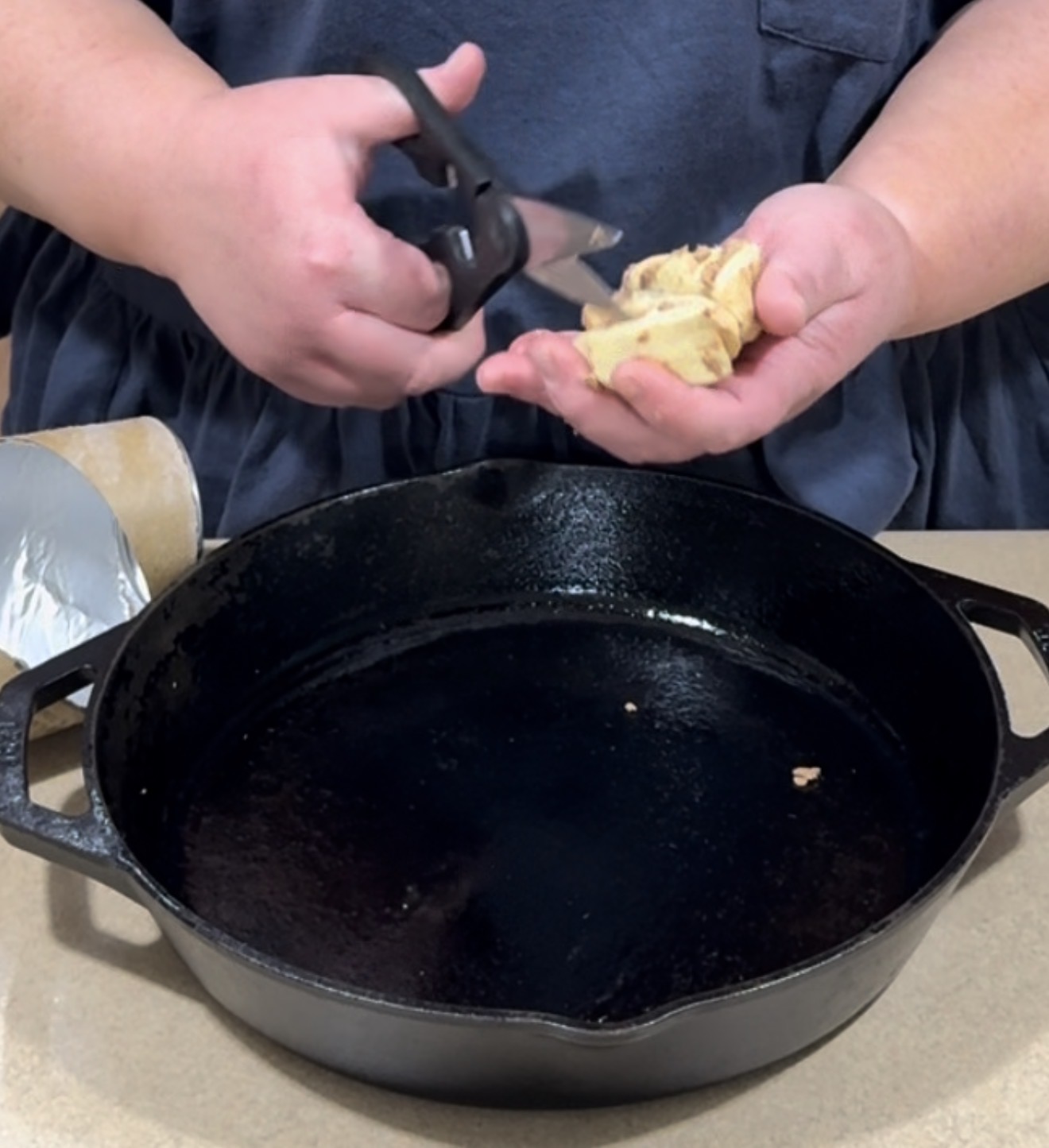 A person cuts pieces of biscuit dough over a black cast iron skillet, preparing to cook. An opened can of biscuit dough is visible on the countertop beside them.