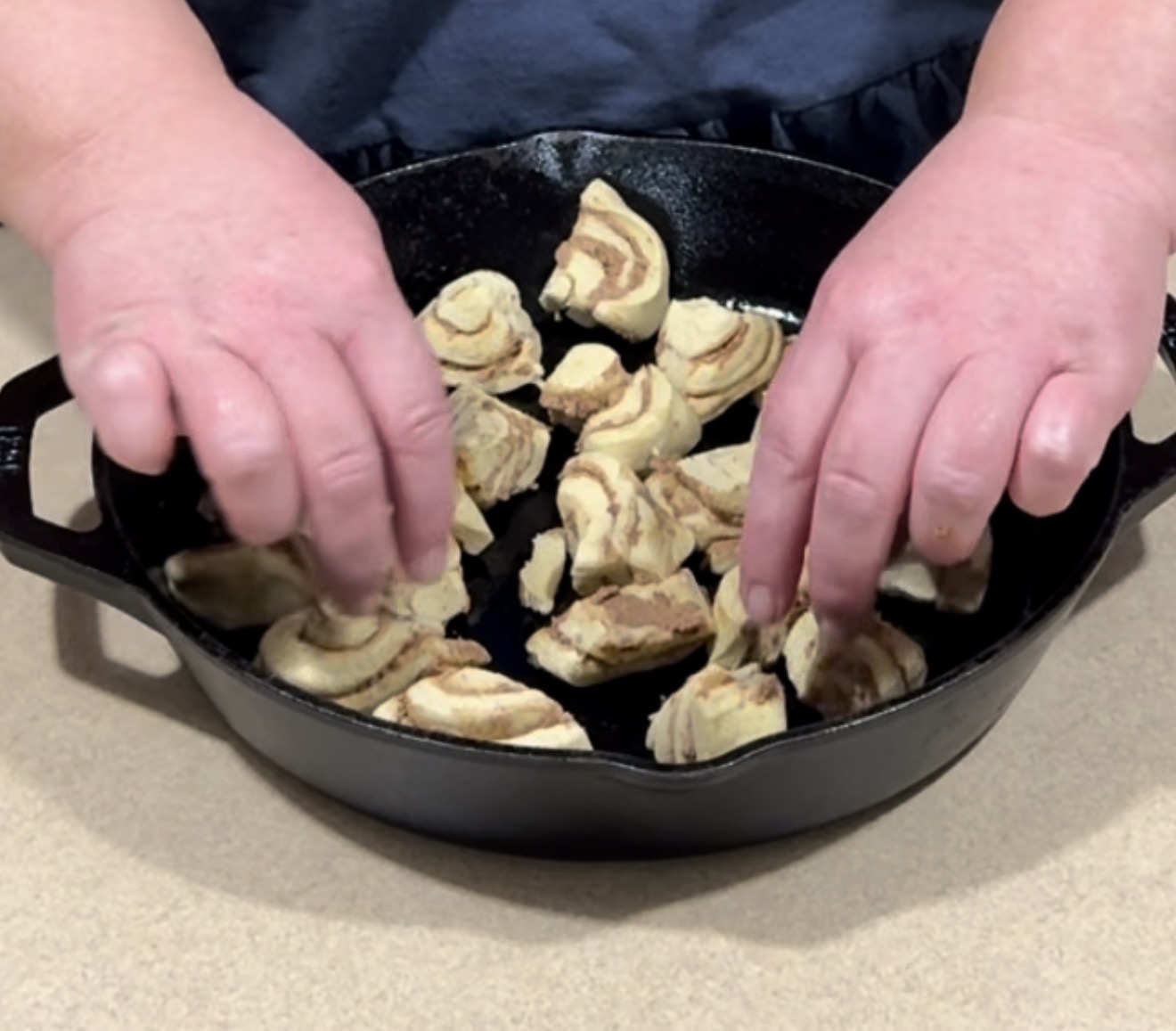 Two hands are arranging pieces of a marbled dough in a black cast iron skillet on a light-colored countertop.