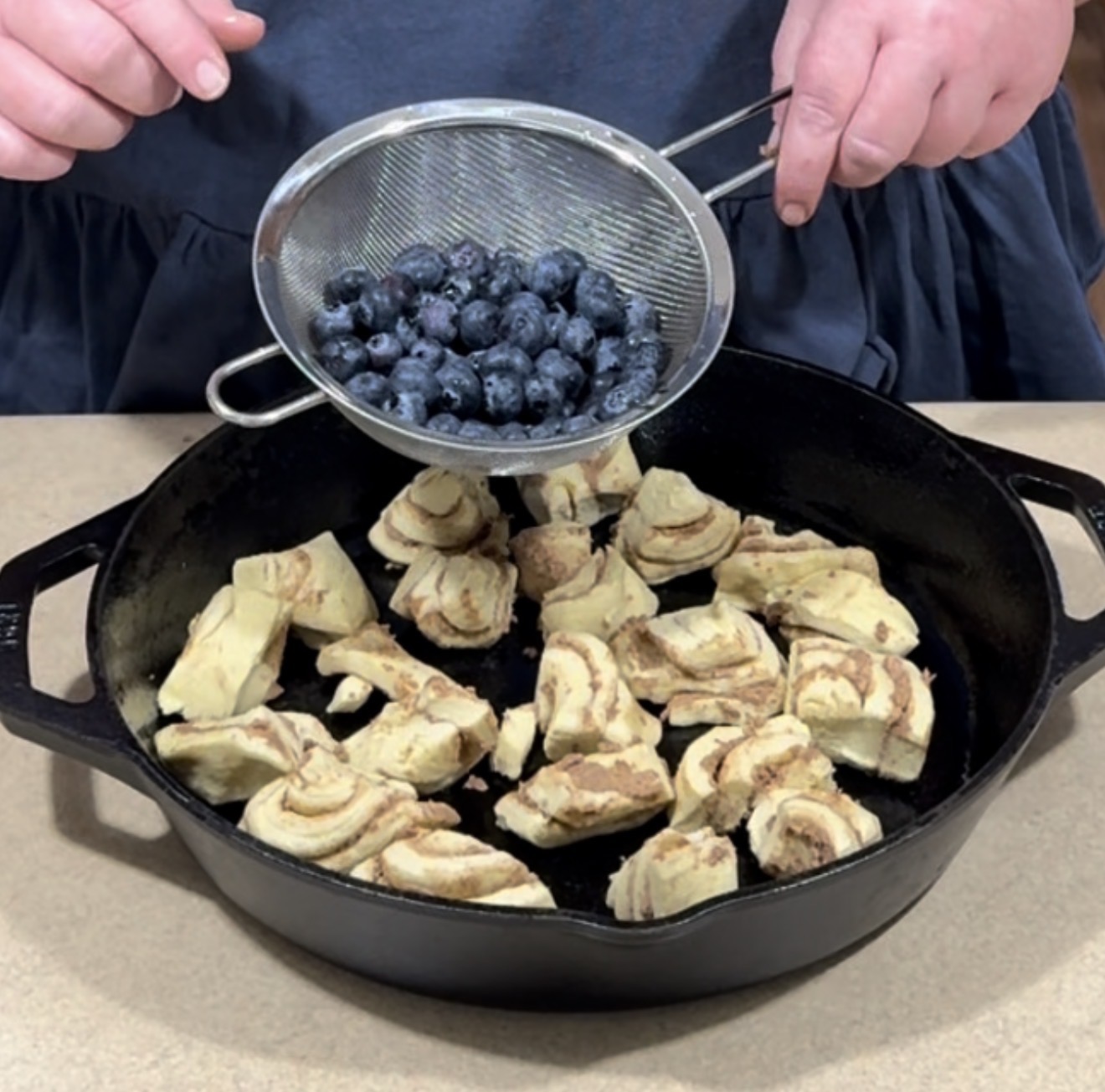 A person holds a strainer filled with fresh blueberries above a cast iron skillet containing pieces of cinnamon rolls, preparing ingredients for baking.