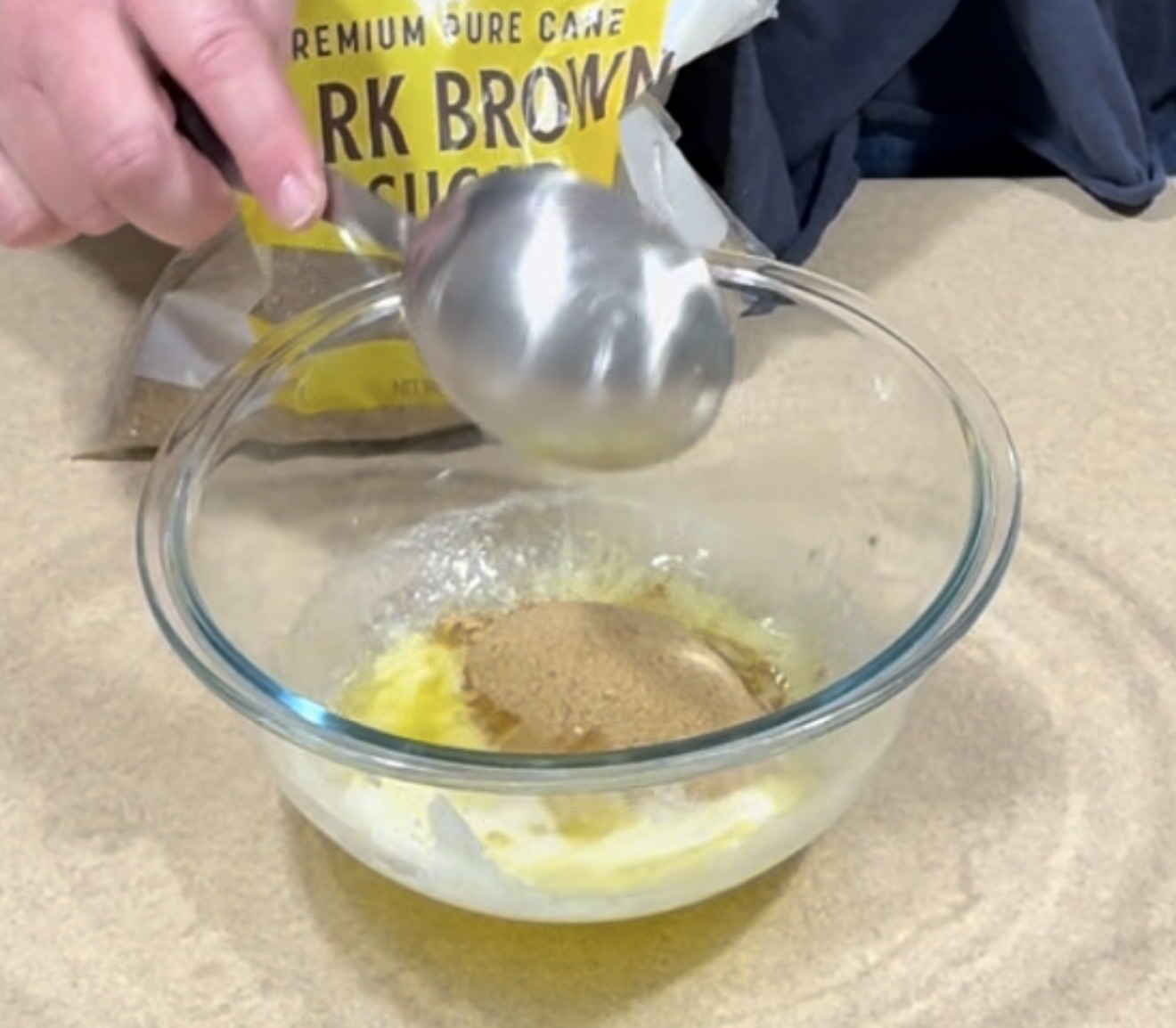 A person holds a metal measuring cup above a glass bowl containing melted butter and brown sugar, with a bag of dark brown sugar in the background on a beige countertop.