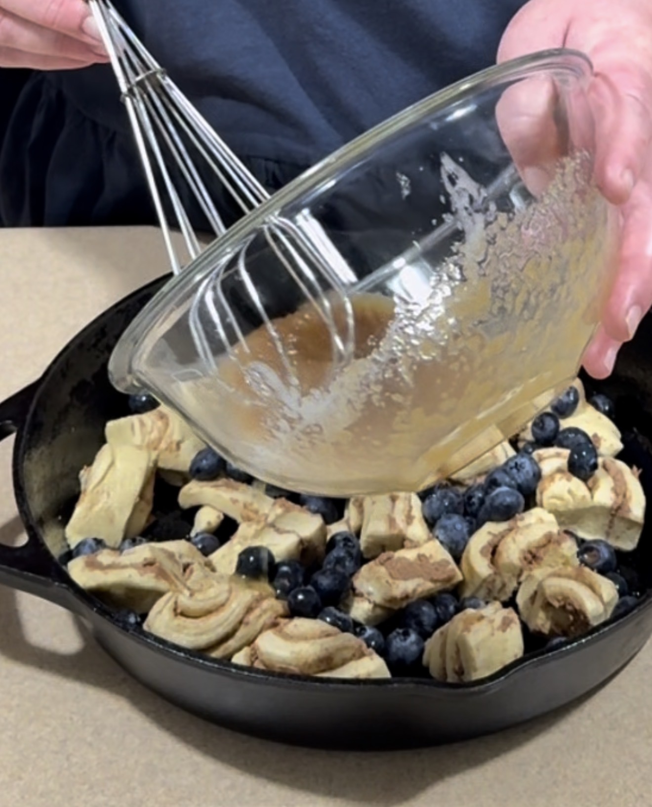 A person pours a liquid mixture from a glass bowl into a cast iron skillet filled with sliced cinnamon rolls and blueberries, preparing a baked dish.