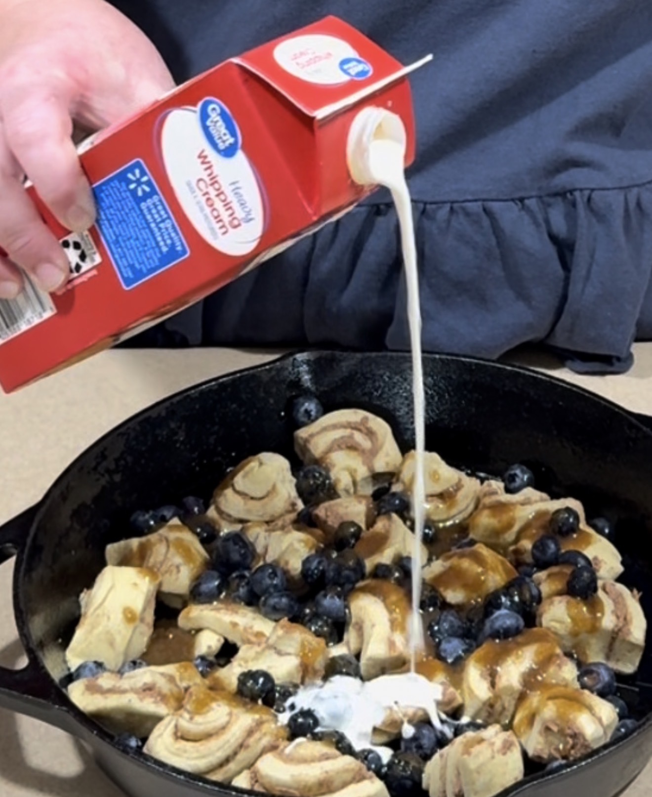 A person pours heavy whipping cream from a carton over chopped cinnamon rolls and blueberries in a black cast iron skillet.