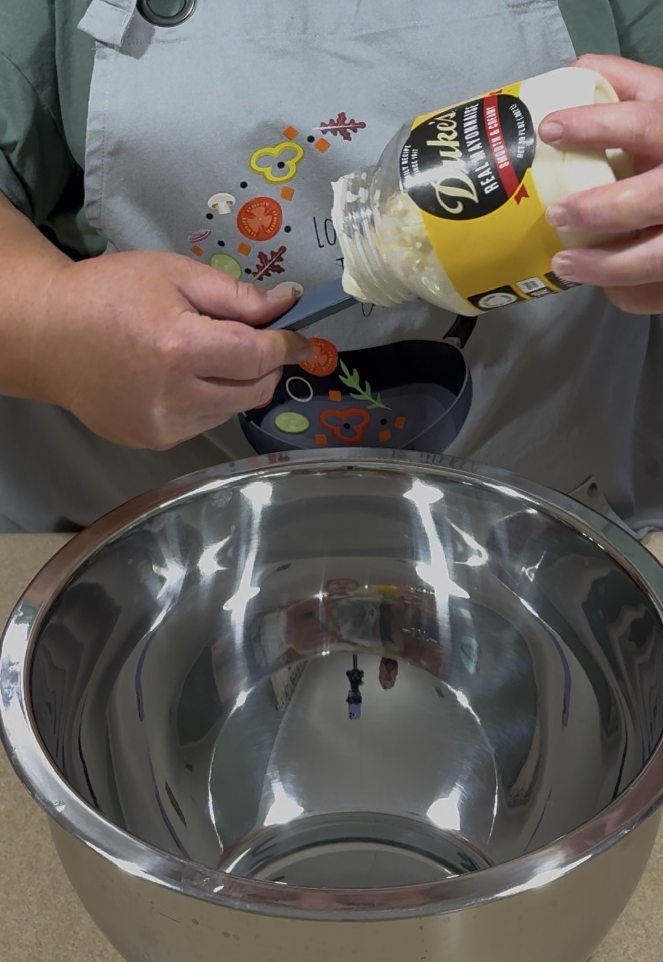 A person in a gray apron is scooping mayonnaise from a jar labeled Dukes into a large, empty stainless steel mixing bowl.