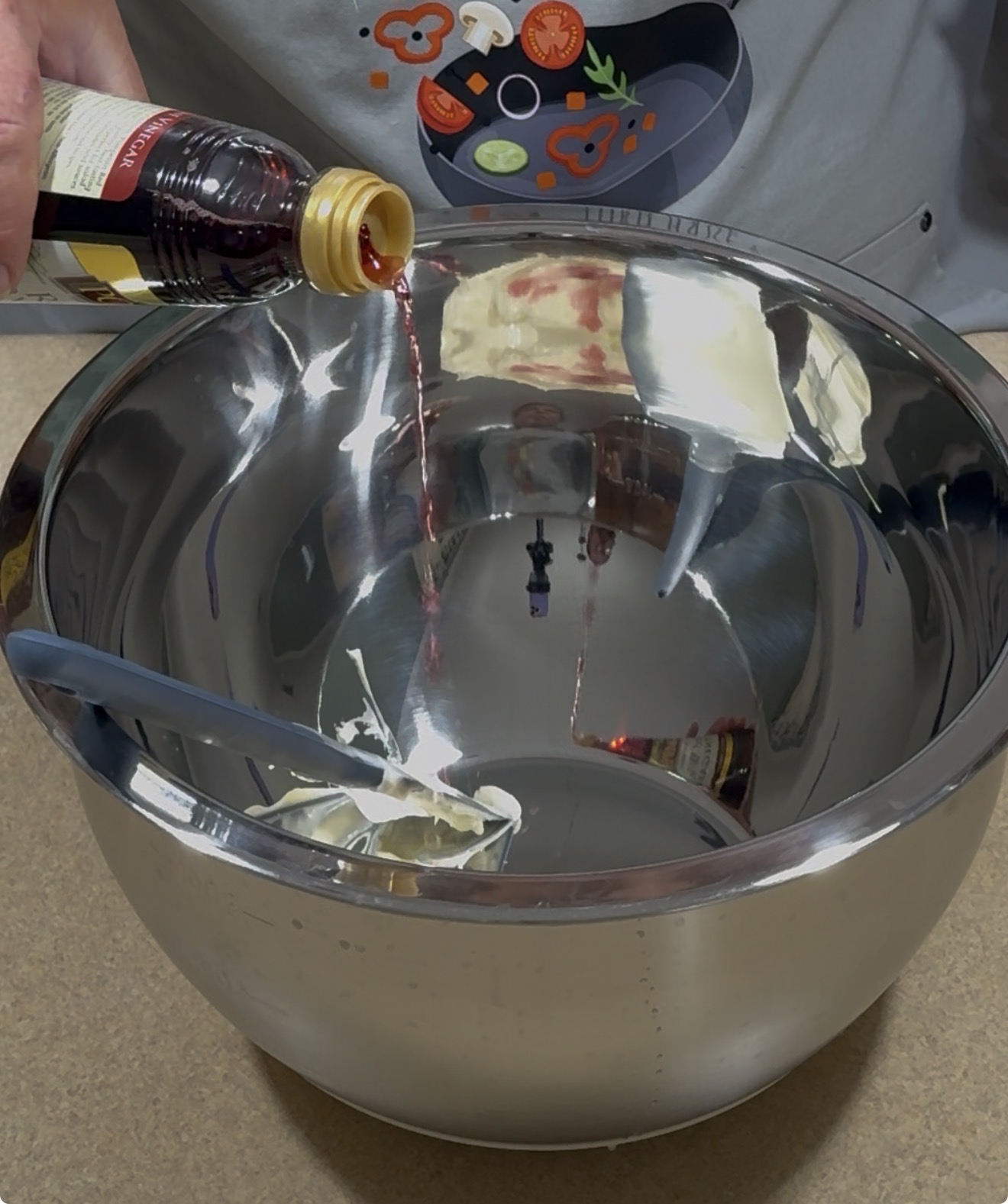 A person pours red liquid from a bottle into a large metal mixing bowl containing a chunk of butter. A spatula rests inside the bowl, and the person is wearing a shirt with colorful food-themed graphics.