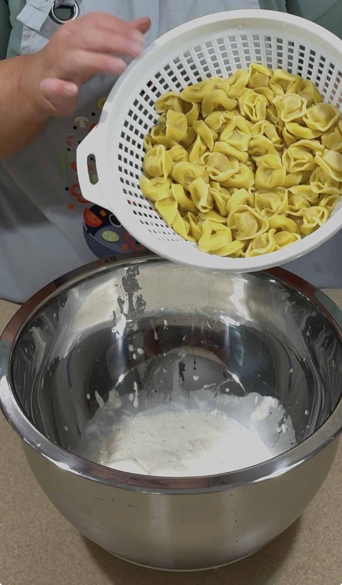 A person pours cooked tortellini from a white colander into a large metal bowl containing a creamy white sauce.