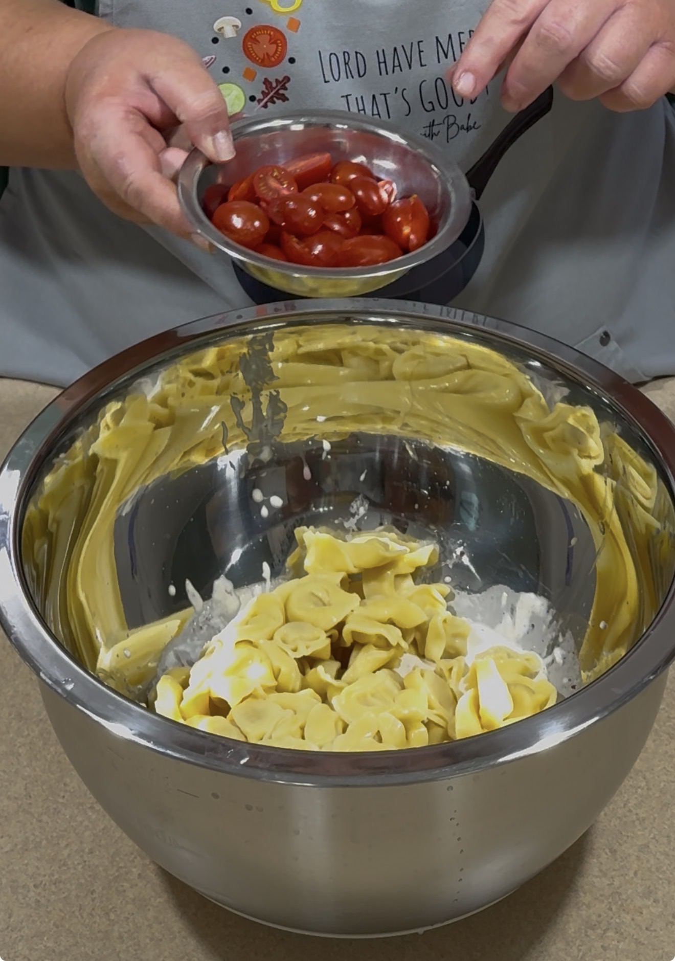 A person adds a bowl of halved cherry tomatoes to a large mixing bowl containing tortellini pasta, mayonnaise, and sour cream. The countertop and part of their shirt are visible in the background.